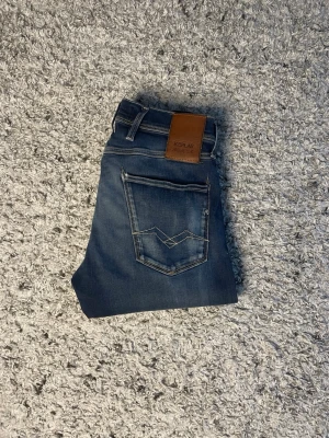 Replay Hyperflex blå jeans - Snygga blå Replay Hyperflex jeans med klassisk femficksdesign och coola slitningar framtill. Jeansen har en skön stretchig känsla och detaljer som kontrastsömmar och läderpatch bak i midjan. Storlek W29 och L32!