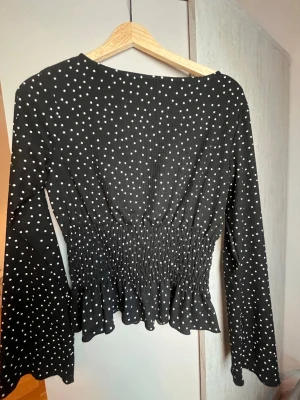 Svart prickig blus Gina Tricot - Snygg svart blus från Gina Tricot med vita prickar. Blusen har lång ärm, smockad midja och volang nedtill för en feminin siluett. Tillverkad i ett mjukt och stretchigt material som sitter bekvämt på kroppen.