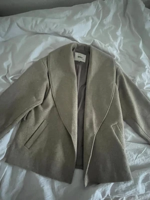 Beige kappa från ONLY med bred krage (har inget snöre till den ) - Stilren beige kappa från ONLY med bred, svepande krage och diskreta fickor framtill. Klassisk rak passform och helfoder, perfekt för dig som gillar minimalistisk och tidlös stil. Passar till många olika outfits.