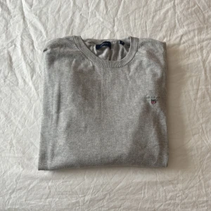 Grå stickad tröja från Gant - Klassisk grå stickad tröja från Gant med rund halsringning och diskret broderad logga på bröstet. Tröjan har långa ärmar och ribbade muddar vid ärmslut och nederkant. Perfekt basplagg i garderoben.