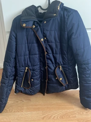 Mörkblå quiltad dunjacka med detaljer - Snygg mörkblå quiltad dunjacka med guldiga dragkedjor och knappar. Jackan har två framfickor med dekorativa detaljer och hög krage. Perfekt för kyliga dagar och ger en clean look.