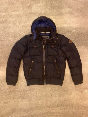 Svart dunjacka från Marc o polo med huva och fickor - Snygg svart dunjacka från Marc o polo med huva, två bröstfickor med knappar och två snedställda fickor med dragkedja. Jackan har quiltad design, ribbade muddar och detaljer i mörkblått foder. Perfekt för kalla dagar. Jackan är i S men funkar också för M. Vid funderingar, fråga på!