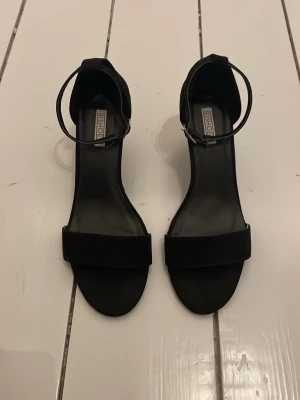 Svarta klackskor  - Stilrena svarta klackskor med öppen tå och smal rem runt ankeln. Skorna är tillverkade i mocka för en mjuk känsla. Perfekta för dig som gillar minimalistisk och klassisk stil.