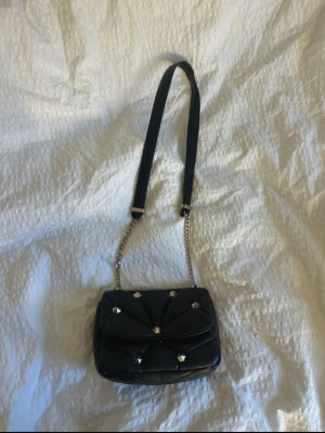 Svart axelväska med nitar - Snygg svart axelväska i quiltad design med coola nitar. Väskan har kedjerem  axelrem i textil. Perfekt storlek för det viktigaste och ger en edgy vibe till din outfit.