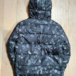 Svart camo pufferjacka från Woolrich - Säljer en svart pufferjacka från Woolrich med grått camouflagemönster. Jackan har huva, dragkedja framtill och är quiltad för extra värme. Perfekt för kalla dagar och har en cool streetstil. Passformen är normal och jackan är långärmad.