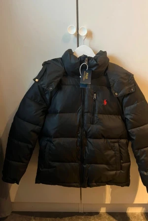 Svart pufferjacka från Polo Ralph Lauren - Svart pufferjacka från Polo Ralph Lauren med röd broderad logga på bröstet. Jackan har huva, dragkedja framtill och flera fickor. Materialet är syntet och jackan är quiltad för extra värme. Perfekt för kalla dagar och har en clean, modern look.