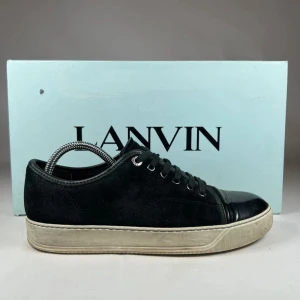 Svarta sneakers från Lanvin - Lanvin sneakers i svart mocka med blankt skinn på tån och snörning. Snygg kontrast mellan den svarta ovandelen och den vita platta sulan. Metallöglor och klassisk rund tå ger en clean och stilren vibe. Perfekta för dig som gillar exklusiva och tidlösa sneakers.