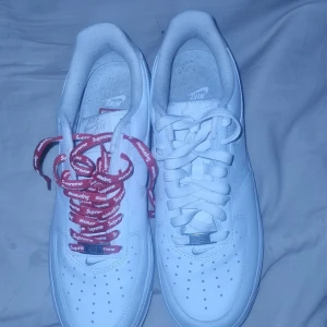Nike Air Force 1 x Supreme vita sneakers - Nike Air Force 1 x Supreme sneakers i helvitt skinn med Supreme-logga på sidan och röda Supreme-skosnören på ena skon. Klassisk låg modell med perforerad tå, platt sula och rund tå. Snygg streetstil och ikonisk design som sticker ut med de röda detaljerna.