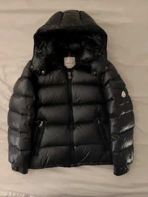 Svart Moncler vinterjacka Maya  - Svart vinterjacka från moncler med en matt glansig färg, går att justera midjemåttet och även ta bort luva för en annan look, parfekt för kyliga dagar. Liten defekt på jackan är att den har ett miniatyr hål vid vänstra armen och en liten sminkfläck, går enkelt att fixa hos en skräddare för billigt. OBS jackan är inte autentisk men håller dig fortfarande varm och stilig under vintern! 