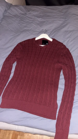 Vinröd kabelstickad tröja - Snygg vinröd kabelstickad tröja med rund hals och långa ärmar. Tröjan har ett klassiskt mönster och ribbade muddar vid ärmslut och nederkant. Perfekt för dig som gillar en chill och stilren look under vinter. Storlek M men passar S.