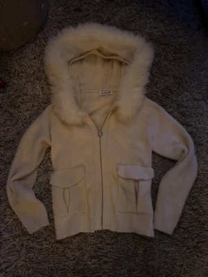 Beige tröja/jacka med fluffig huva - Mysig beige tröja från Bluoltre med fluffig fuskpäls runt huvan. Jackan har två stora fickor framtill och stängs med dragkedja. Perfekt för dig som gillar en chill och trendig look med extra cozy vibes.