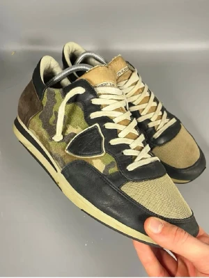 Philippe Model sneakers med camo - Storlek 45 men passar även 44 .Snygga sneakers från Philippe Model med coolt kamouflagemönster i grönt och brunt, svarta skinnpaneler och beige mesh. Skorna har vita snören och detaljer i mocka och läder. Perfekta för dig som vill sticka ut med en urban och sportig stil.