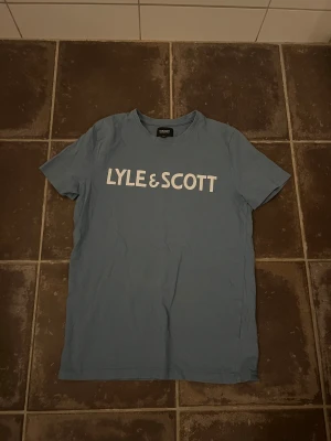 Lyle & Scott t shirt - Snygg blå t-shirt från Lyle & Scott med stort vitt logotryck på bröstet. Klassisk rund hals och korta ärmar. Tillverkad i mjuk bomull som är skön mot huden. Perfekt för dig som gillar stilrena och enkla plagg med tydlig branding.