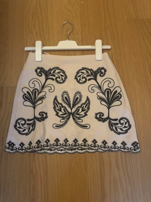 Vit kortkjol med svart broderi - Supersnygg vit kortkjol med svart broderat mönster och dekorativ kant längst ner. Kjolen har en rak passform och är perfekt för dig som gillar detaljer och kontraster. Broderiet ger kjolen en unik och cool vibe.