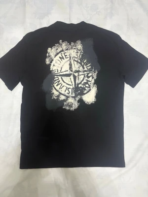 Stone Island Tröja - Svart t-shirt från Stone Island med stort grafiskt tryck i vitt på ryggen och klassisk logotyp på bröstet. Modellen har rund hals och korta ärmar. Materialet är mjuk bomull som känns skön mot huden. Perfekt för dig som gillar streetwear och vill sticka ut.