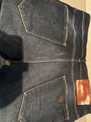 Emporio Armani mörkblå jeans - Snygga mörkblå jeans från Emporio Armani med klassisk femficksdesign och kontrastsömmar. Byxorna har ett stilrent läderpatch med logga bak och metallknapp framtill. Perfekta för dig som vill ha en clean och tidlös look.