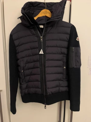 Svart Moncler pufferjacka med huva - Snygg svart pufferjacka från Moncler med stickade ärmar och vadderad kropp. Jackan har huva, dragkedja framtill, fickor med tryckknapp och en extra ficka på ärmen. Perfekt för kyliga dagar och har en clean, modern look.