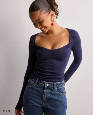 Mörkblå långärmad topp med v-ringning - Snygg mörkblå långärmad topp med djup v-ringning och diskret spetskant runt halsen. Figurnära passform som framhäver siluetten. Tillverkad i ett stretchigt och mjukt material som känns skönt mot huden. Perfekt att styla med jeans eller kjol.