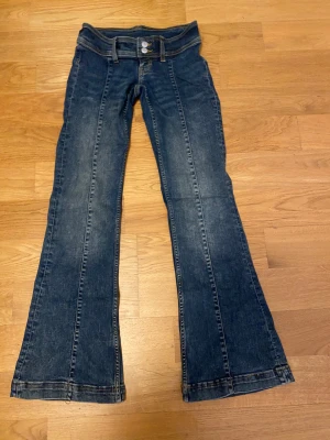 Blå bootcut jeans med låg midja 25/32 - Snygga blå jeans med bootcut och låg midja, klassisk femficksmodell och dubbla silverknappar fram. Jeansen har kontrastsömmar och är tillverkade i slitstarkt denim. Perfekt passform för dig som gillar retrovibbar och markerad siluett.