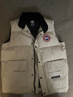Vit dunväst från Canada Goose - Säljer en vit dunväst från Canada Goose med svart krage och klassisk logga på bröstet. Västen har två stora fickor framtill, dragkedja och tryckknappar. Insidan är svart och har meshficka. Perfekt för kalla dagar när du vill ha en clean och stilren look.