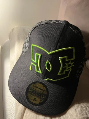DC keps - Säljer en svart DC x New Era 59FIFTY keps i storlek S, med stort limegrönt DC-logo broderat framtill och mönstrade och glimrande paneler på sidorna. Kepsen är tillverkad i 100% akryl och har en klassisk flat brim. Perfekt för dig som gillar streetwear och skate.