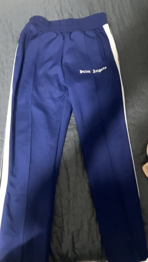 Palm Angels blå trackpants med vita ränder - Snygga blå trackpants från Palm Angels med vita sidoränder och broderad logga på framsidan. Byxorna har elastisk midja med vit snörning och är tillverkade i ett mjukt, syntetiskt material. Perfekt för en sportig och trendig look.