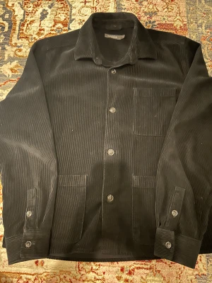 Svart manchester overshirt Dressmann - Svart overshirt från Dressmann i mjuk manchester. Klassisk krage, tre fickor fram och knäppning med knappar. Långärmad modell med knappslå vid ärmslut. Perfekt för lager-på-lager och chill stil.