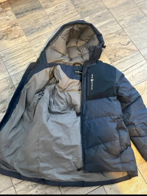 Mörkblå pufferjacka från Sail Racing - Säljer en mörkblå pufferjacka från Sail Racing med huva och grått foder. Jackan har en dragkedja framtill och en praktisk bröstficka med logga. Perfekt för kalla dagar och har en schysst, lite oversized passform.