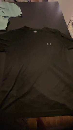 Svart t-shirt från Under Armour - Svart t-shirt från Under Armour med klassisk rund hals och diskret logga på bröstet. T-shirten är kortärmad och har en clean, sportig look som passar perfekt till gymmet eller chill dagar. Materialet är mjukt och lätt, vilket gör den skön att bära. Pris kan diskuteras!