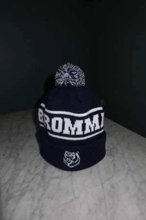 Bromma Mössa - Snygg mörkblå mössa med vit text 'BROMMA' och broderad björn från SkiStyle UF. Perfekt för dig som vill visa Bromma-stolthet och hålla dig varm, skriv för frågor eller funderingar! 