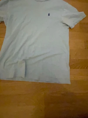 Vit t-shirt från Polo Ralph Lauren - Vit t-shirt från Polo Ralph Lauren med klassisk blå broderad logga på bröstet. Modellen är enkel med rund hals och korta ärmar, perfekt för en clean och stilren look. Materialet är mjuk bomull som känns skönt mot huden.