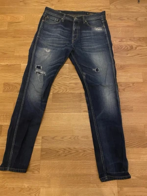 Jack & Jones mörkblå jeans 32/32 - Snygga mörkblå jeans från Jack & Jones Originals, modell Erik Original Felling Piping. Jeansen har slitna detaljer och hål på framsidan för en cool, avslappnad look. Klassisk femficksdesign med kontrastsömmar och raka ben.