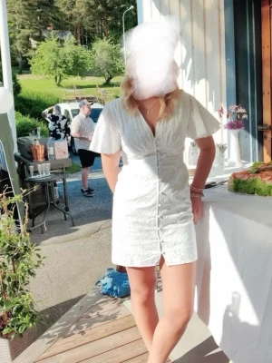 Vit kort klänning från NA-KD - Supersöt vit klänning från NA-KD med broderade detaljer och knappar framtill. Klänningen har v-ringning, korta ärmar och en figurnära passform. Perfekt för sommarens alla tillfällen! Materialet är bomull och ger en luftig känsla.