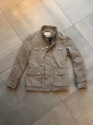 Grå field jacket från Zara Man - Säljer nu denna field jacka från Zara Man med ståkrage och dragkedja framtill. Fyra fickor med knappar på framsidan och justerbara ärmslut med knappdetaljer. Stolek:(S) Skick:(9/10). Säljer då den inte längre passar mig