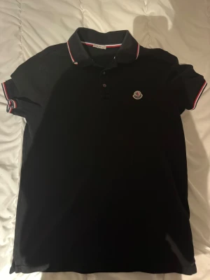 Svart Moncler piké med röd/vit kant - Svart pikétröja från Moncler med klassisk krage och korta ärmar. Röd och vit randig kant på krage och ärmslut, samt Moncler-logga på bröstet. Tröjan har knappar framtill och är gjord i mjuk bomull som känns skön mot huden. 