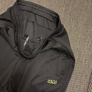 Svarta träningsbyxor från asos - Svarta träningsbyxor från asos med smal passform och elastisk midja med snörning. Byxorna har sidofickor och en diskret logga i gult på benet. Perfekta för gymmet eller chill hemma, tillverkade i mjukt syntetmaterial. Storlek S men passa M också