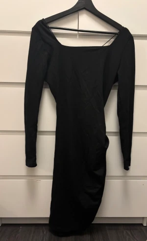 Svart klänning från NA-KD  - Svart klänning från NA-KD i mjuk bomullsmix. Klänningen har långa ärmar, snygg v-ringning med snörningsdetalj baktill och drapering längs ena sidan för en cool siluett. Perfekt för dig som gillar stilrena och trendiga plagg.