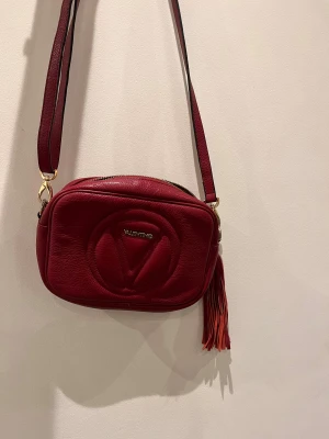 Röd axelväska från Valentino - Snygg röd axelväska från Valentino med guldiga detaljer och en cool fransad tofs på sidan. Väskan har ett präglat V på framsidan och dragkedja upptill. Avtagbar axelremband