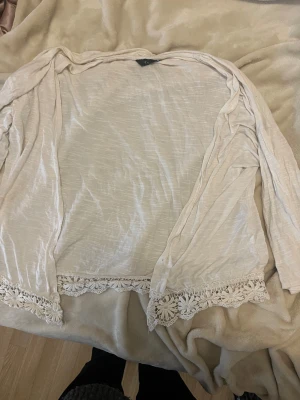 Beige kofta med spetsdetaljer - Säljer en tunn och lätt beige kofta från Vero Moda med långa ärmar. Koftan har snygga spetsdetaljer längs nederkanten som ger en boho-vibe. Perfekt att slänga över en topp när du vill ha en chill och stilren look.