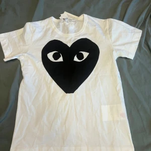 Vit t-shirt Comme des Garçons Play  - Snygg vit t-shirt från Comme des Garçons Play i storlek S. Framsidan har ett stort svart hjärta med ögon, vilket ger en unik och cool vibe. Klassisk rund hals och korta ärmar. Tillverkad i mjuk bomull för skön känsla hela dagen. Pris ej hugget i sten!
