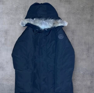 Svart Canada Goose parkas med päls - Säljer en svart parkas från Canada Goose med huva och snygg pälsdetalj. Jackan har ett broderat märke på ärmen och dragkedja framtill. Perfekt för kalla vinterdagar och har en klassisk, clean look. Medföljer originalpåse från Canada Goose.