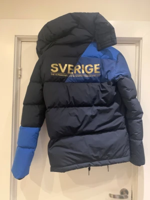 Blå pufferjacka Sverige Paralympics H&M - Säljer en fet pufferjacka från H&M, designad för Sveriges Paralympics-trupp 2018. Jackan är mörkblå med ljusblå detaljer och har guldtryck med SVERIGE och SWE på ryggen och bröstet. Stor huva, dragkedja och snörning nedtill. Perfekt för kalla vinterdagar.