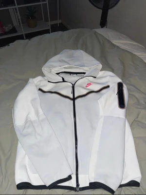 Vit hoodie från Nike med dragkedja - Säljer en vit hoodie från Nike i storlek M. Tröjan har svart dragkedja, huva och svarta detaljer vid muddar och nederkant. På bröstet finns en rosa Nike-logga och en svart vågig linje. Långärmad med tumgrepp och ficka på ärmen. Perfekt för chill eller träning. Ett hål i längst ner i armen därav mitt pris! Skriv om det finns frågor!