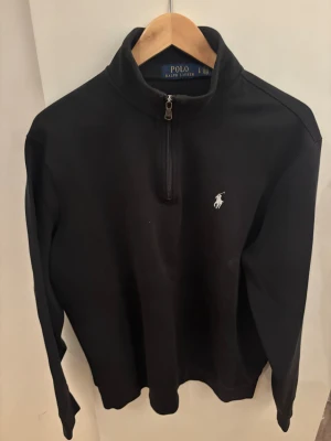 Svart half zip tröja Polo Ralph Lauren - Svart långärmad tröja från Polo Ralph Lauren med half zip och klassisk vit broderad logga på bröstet. Tröjan har en hög krage och är tillverkad i mjuk bomull, perfekt för en chill och stilren look.