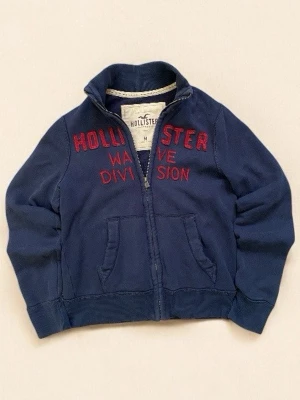 Vintage Navy Hollister Full-Zip - Snygg marinblå ziptröja från Hollister med röd broderad text på bröstet. Tröjan har ståkrage, två fickor fram och dragkedja hela vägen. Mjuk bomullskvalitet som är skön att ha på sig. Liten skada på framfickan, men det tycker jag bara lägger till mer till stilen