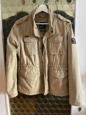 Safari Jacka - Superdry  - Snygg beige parkajacka från Superdry med fyra stora fickor framtill, coola axelklaffar och hög krage med dragkedjedetalj. Jackan har knappar och justerbar midja för skön passform. Perfekt för dig som gillar streetstyle och funktion.