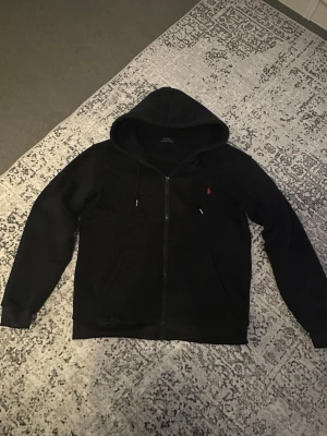 Polo Ralph Lauren Zip-Hoodie - Säljer en klassisk svart Polo hoodie i nytt skick. Tröjan är gjord i mjukt och kvalitativt material som både sitter bra och känns skön att bära. Passar perfekt till både vardag och mer uppklädda outfits.