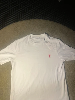Vit t-shirt från Ami med rött hjärta - Vit t-shirt från Ami Alexandre Mattiussi med klassisk rund hals och korta ärmar. På bröstet finns ett broderat rött hjärta med bokstaven A. Materialet är mjuk bomull och baktill syns en diskret AMI-brodering vid nacken. En clean och stilren tee med snygga detaljer.