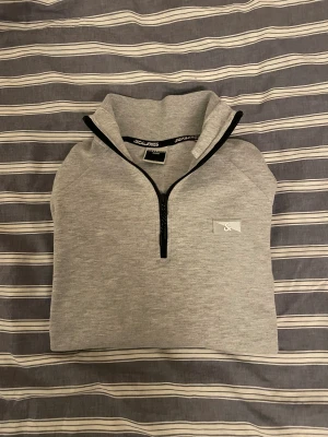 Jack & Jones Quater Zip  - Snygg grå långärmad tröja från Jack & Jones med svart quater zip-dragkedja och liten logga på bröstet. Tröjan har en hög krage och svarta detaljer vid dragkedja och nederkant. Perfekt för chill eller när du vill vara casual.