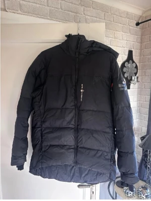 Sail racing jacka - Svart pufferjacka från Ski Patrol med huva och dragkedja framtill. Jackan har broderad logga på ärmen och detaljer på huvan. Perfekt för kalla dagar med sin tjocka, quiltade design och långa ärmar. Snygg och funktionell för vinterväder.